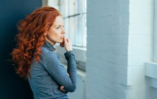 Sad lonely thoughtful young woman with gorgeous long curly red hair standing sideways indoors staring though a window