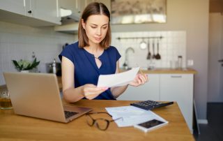 Check Your CSA Income Estimate. Accompanying image: thoughtful young woman using a laptop computer sitting at her kitchen holding utility bill and bank statements.
